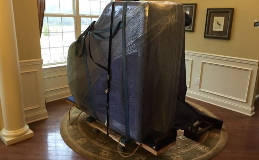 A Piano Moving Company safely transports a grand piano wrapped in protective padding and secured with straps inside a home.