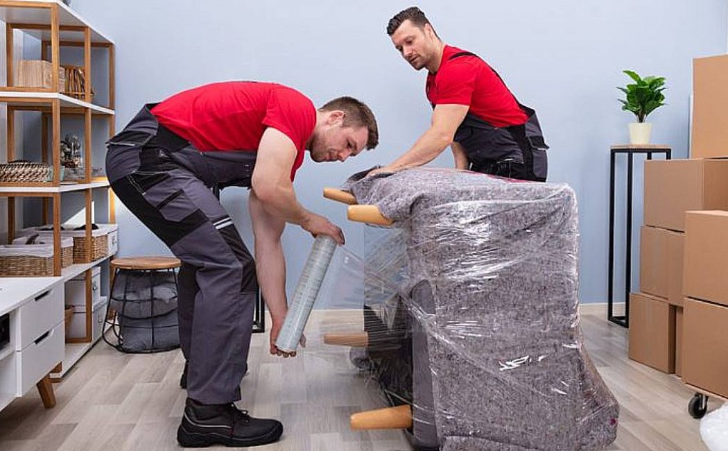 Two professional movers wrapping and securing furniture in a living room with packed boxes, representing Residential Moving Services.