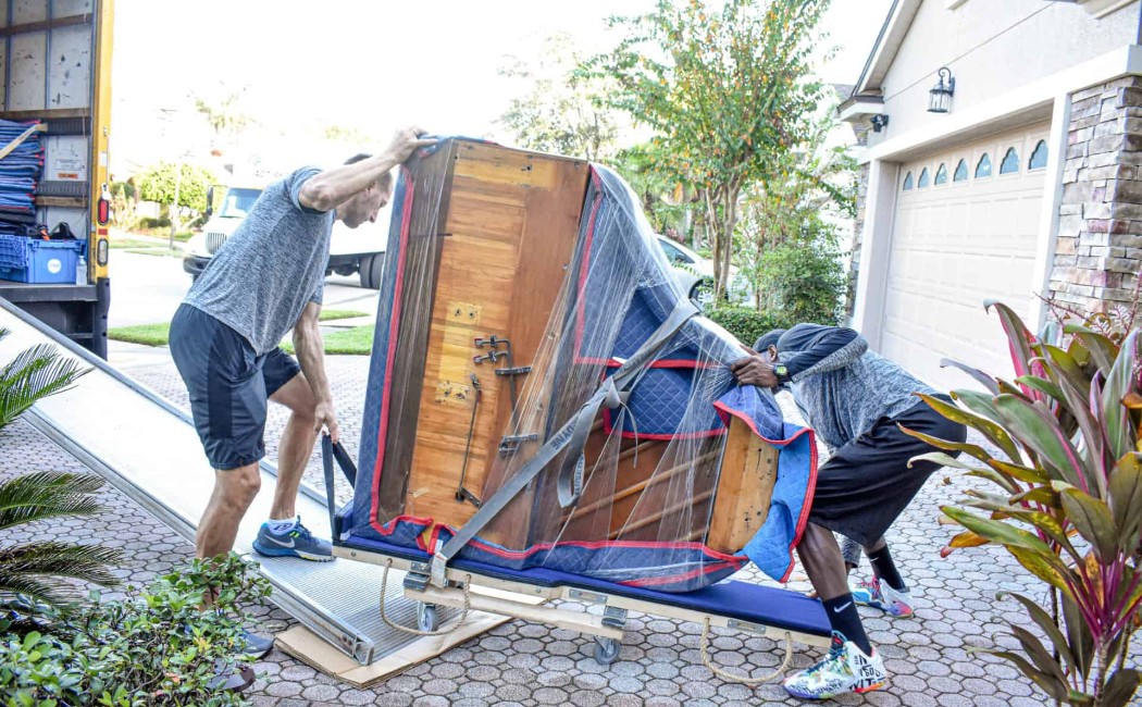Two movers carefully handling and transporting a wrapped upright piano down a ramp into a truck during a piano moving job.