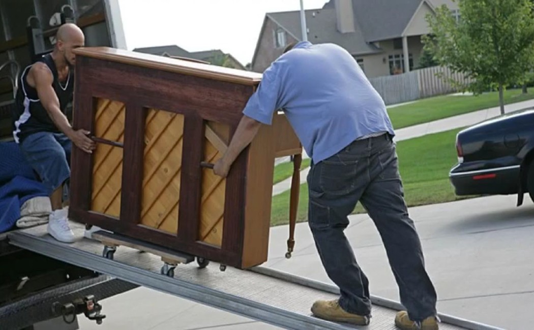 Two movers carefully unloading an upright piano down a ramp from a truck in a residential area during a piano moving service.