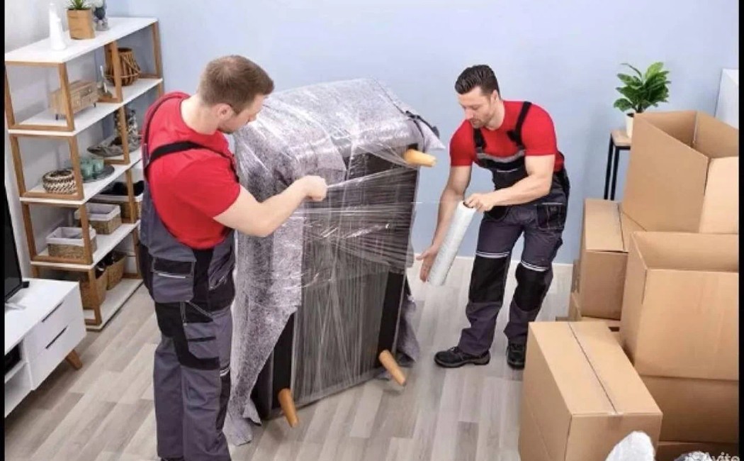 Two movers wrapping furniture in protective film amid boxes, demonstrating professional Packing and Unpacking Services in a modern home.